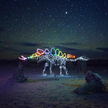 Glowing stegosaurus made of light in a desert landscape with stars and the Milky Way above, captured in a long-exposure photograph