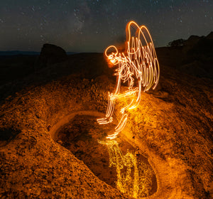 An archangel skeleton with glowing wings created using light painting photography, sitting on a rock with a reflection in the water.