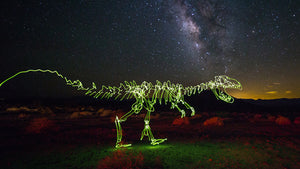 A cosmic T-rex dinosaur drawn with light painting beneath the Milky Way during a long-exposure night photograph.
